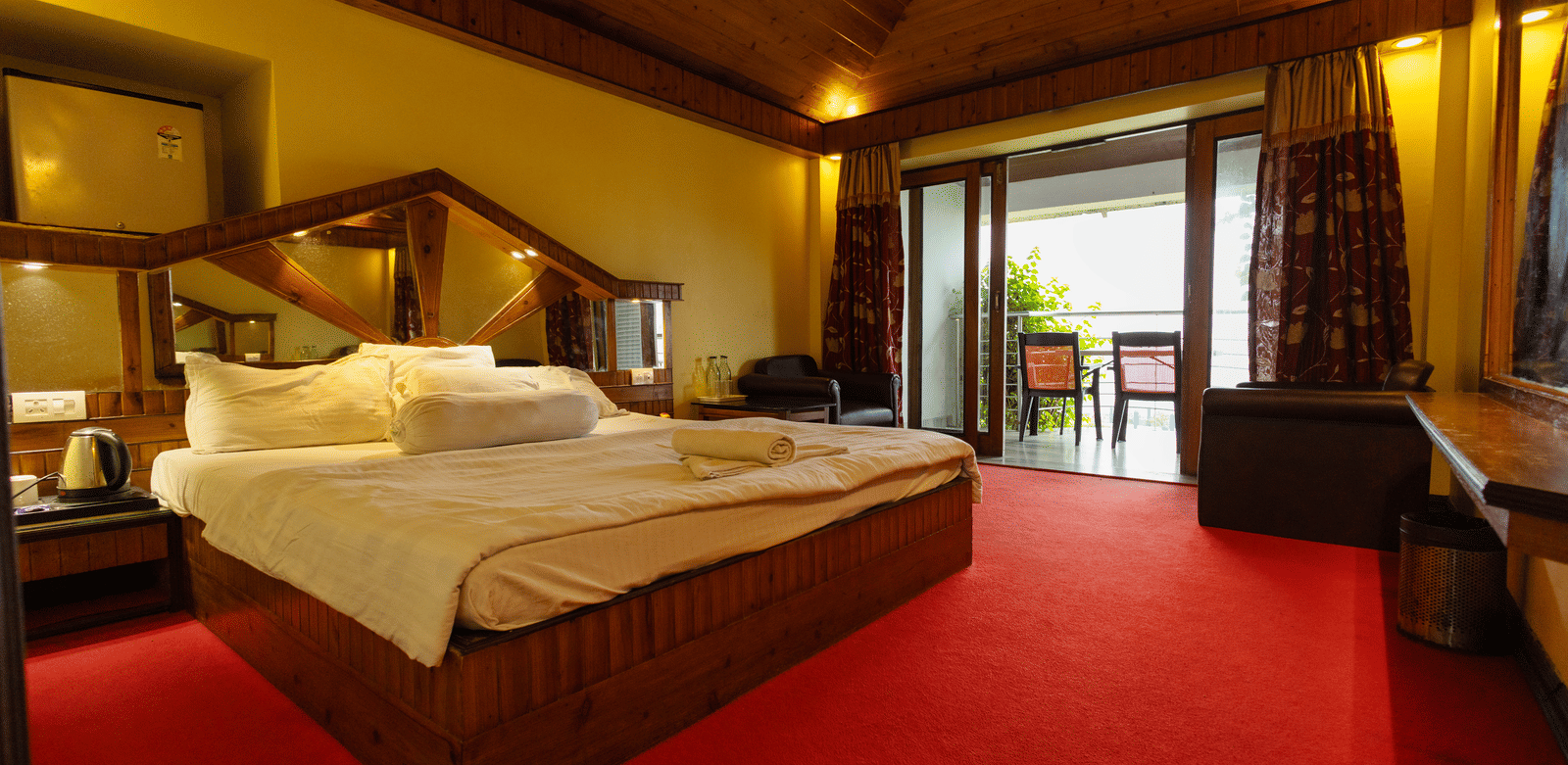 A bedroom with a king size bed, seating area, red carpet, and open balcony door at Hotel Seagull Digha.